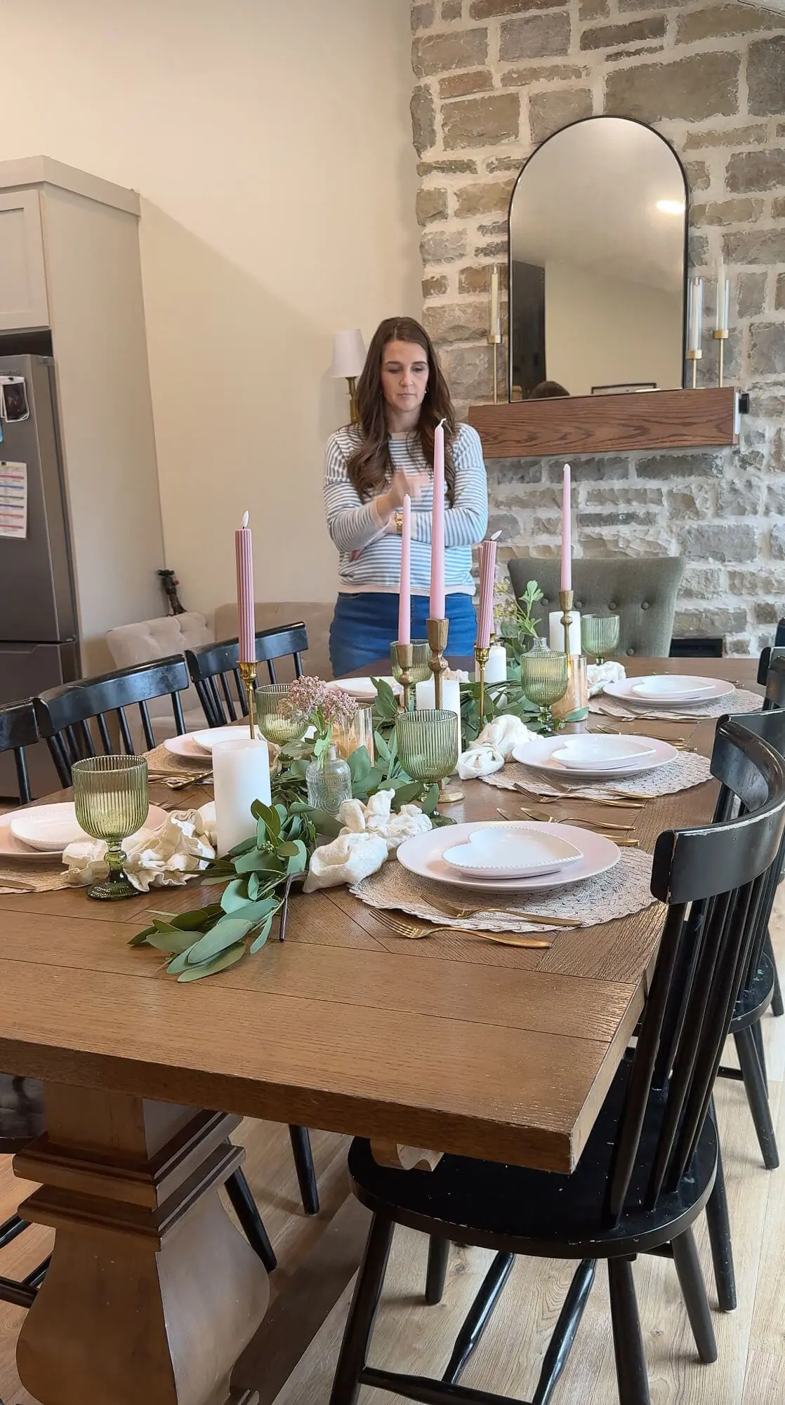 A completed dining table styled for a romantic setting, showcasing pink dinner plates, white heart-shaped dishes, green goblets, and gold candlesticks with pink taper candles. A woman stands at the end of the table, making final adjustments to the decor.